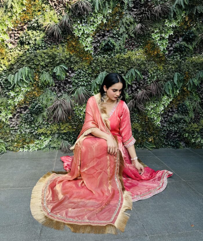 A woman wearing a pink Tissue Silk Anarkali suit sitting in front of a stylish wall backdrop.