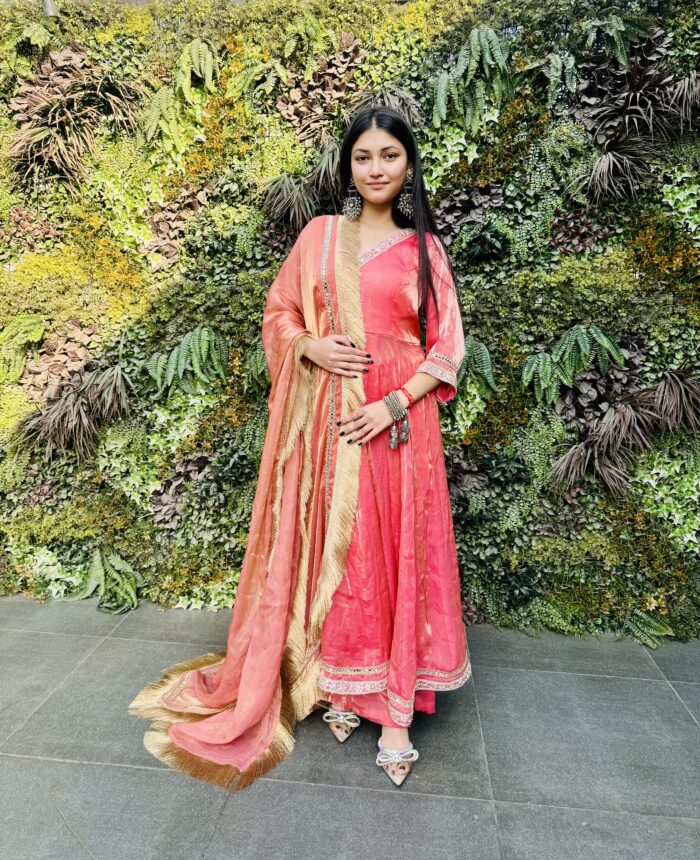 A woman wearing a pink Tissue Silk Anarkali suit poses in front of a stylish wall backdrop.